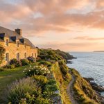 Une maison en pierre traditionnelle au bord d'une falaise surplombant la mer au coucher du soleil.